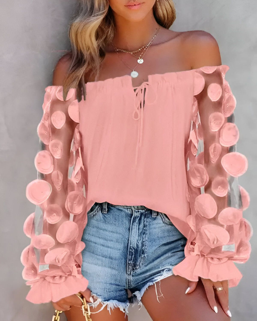 Une jeune femme porte un chemisier rose à épaules dénudées. Le chemisier est en mousseline de soie et comporte des manches bouffantes. La femme est également vêtue d'un short en jean bleu et de sandales roses.