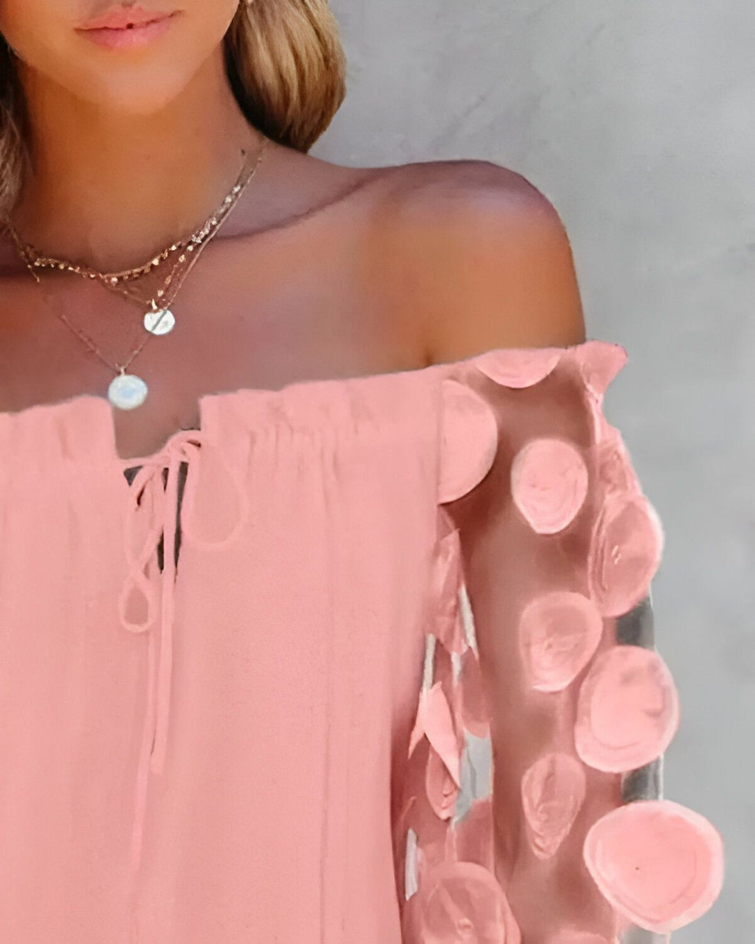 Une jeune femme porte une robe rose à épaules dénudées. La robe est en mousseline de soie et comporte des détails floraux sur les manches. La femme a les cheveux blonds et porte un collier.