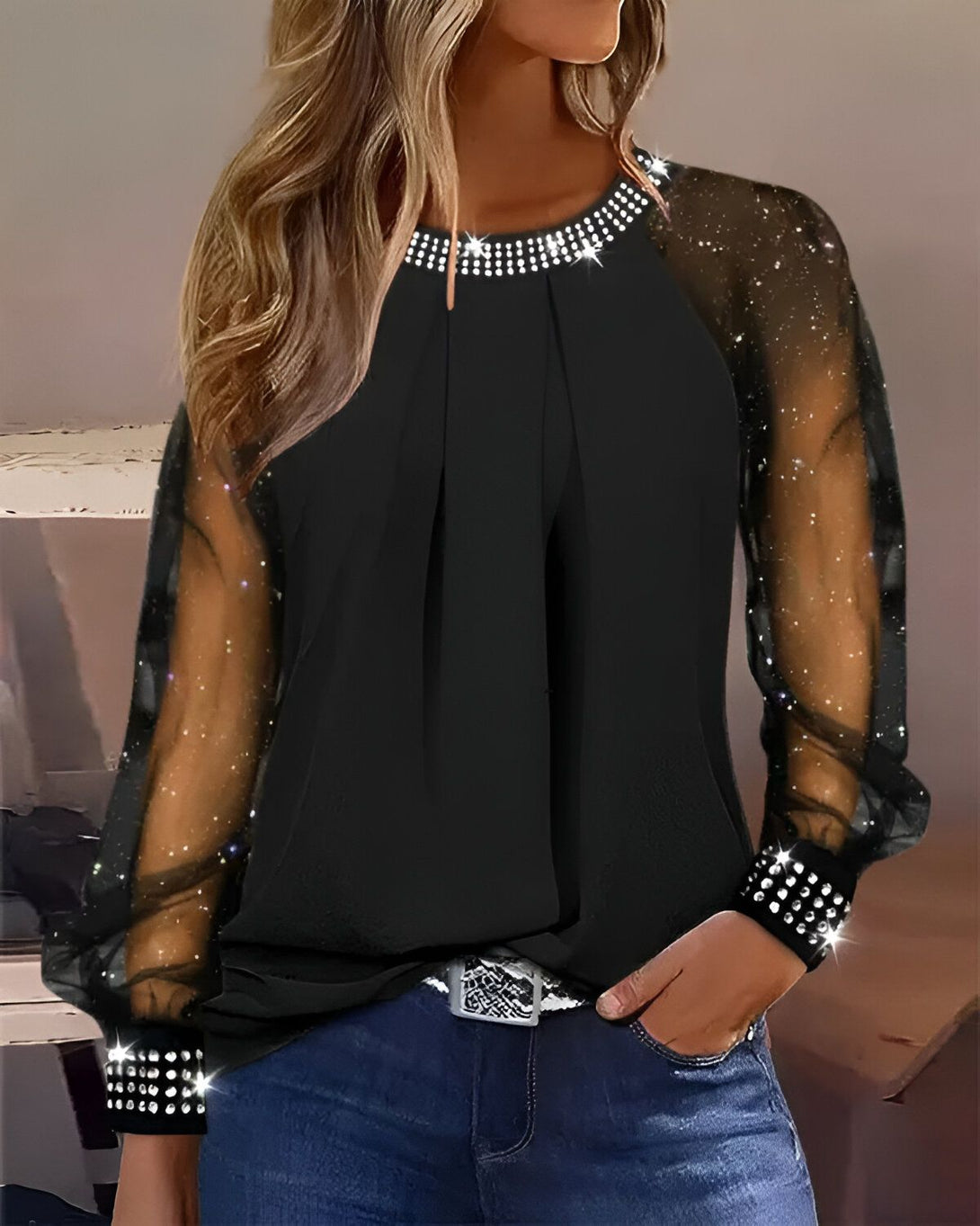 Une femme porte un chemisier noir à manches longues en mousseline transparente parsemée de paillettes argentées. Le chemisier est orné de strass au niveau du col et des poignets.