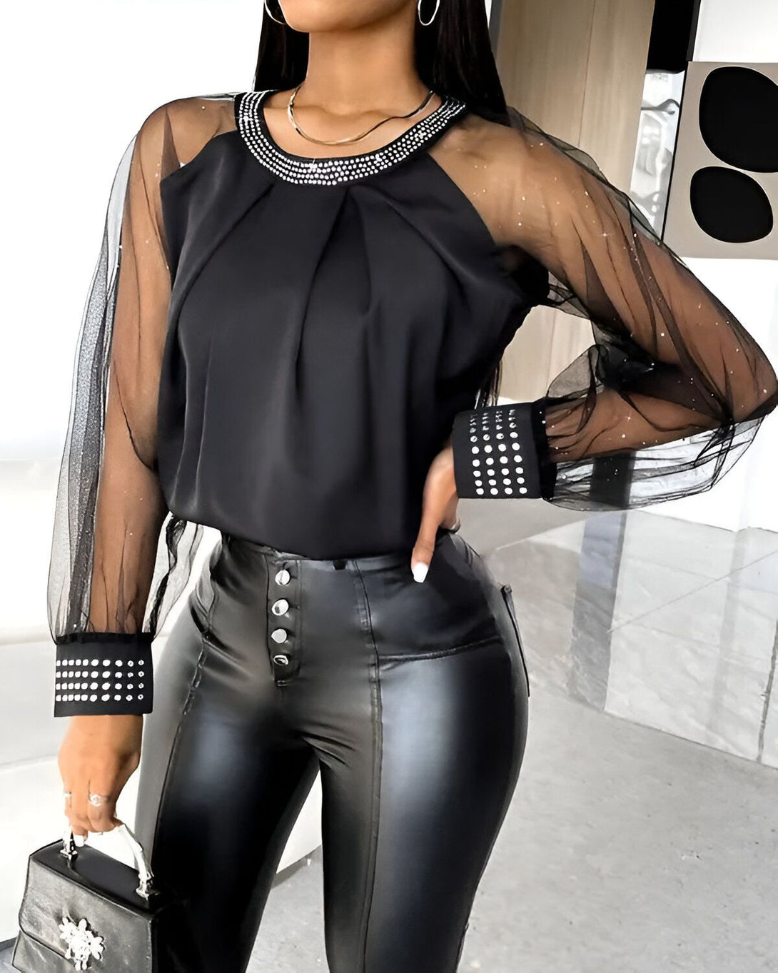 Une femme porte un chemisier noir transparent à manches longues avec des détails en strass. Elle porte également un pantalon en cuir noir et un collier.