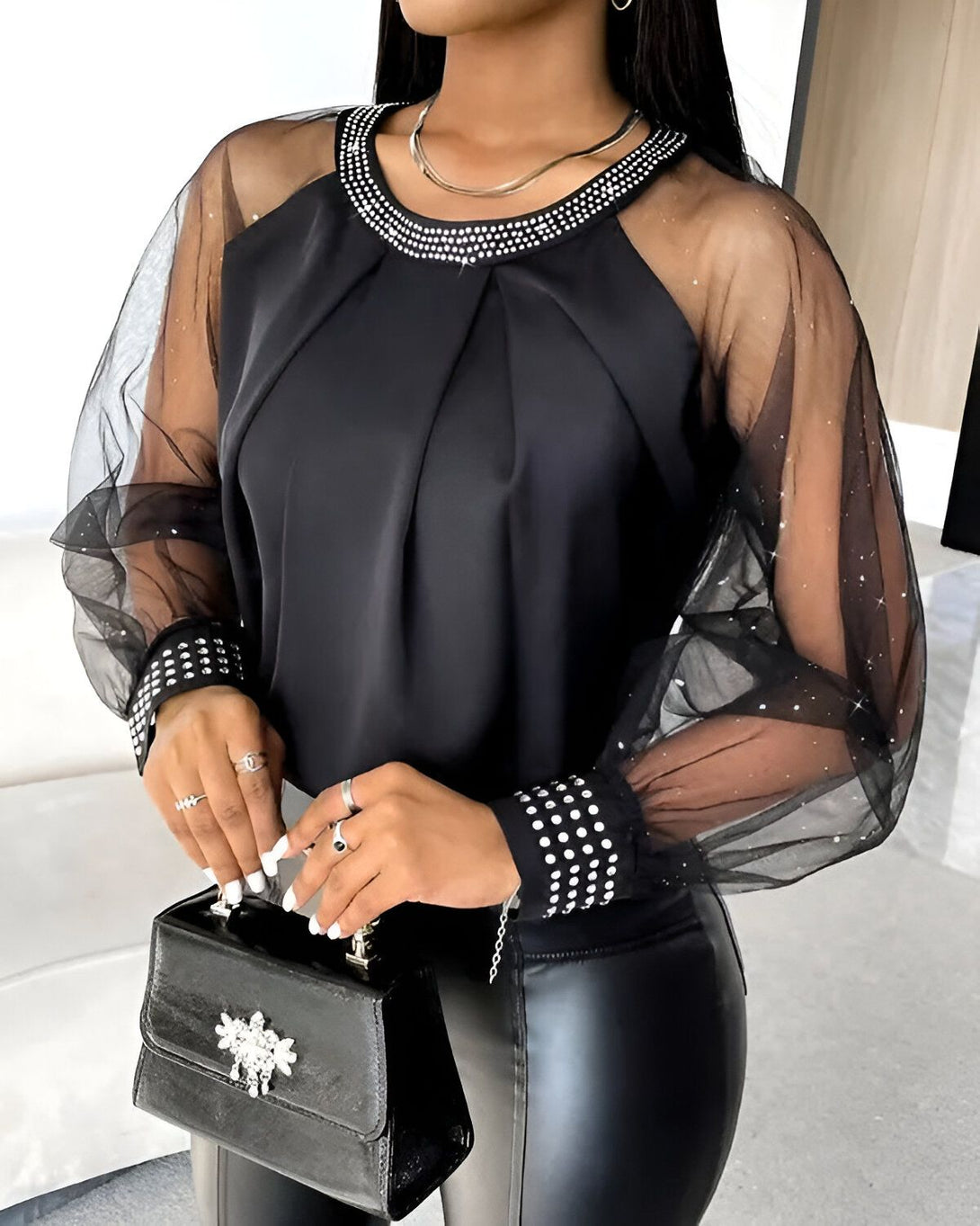 Une femme porte un chemisier noir transparent à manches longues. Le chemisier est orné de strass au niveau du col et des poignets. La femme porte également un pantalon en cuir noir et des escarpins noirs.