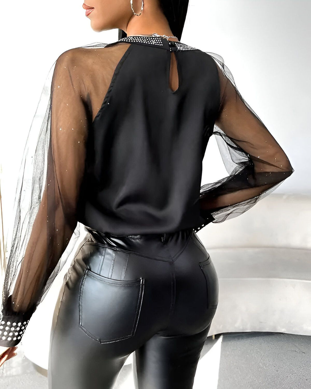 Une femme porte un chemisier noir transparent à manches longues avec des strass au niveau du col et des poignets. Elle porte également un pantalon en cuir noir.