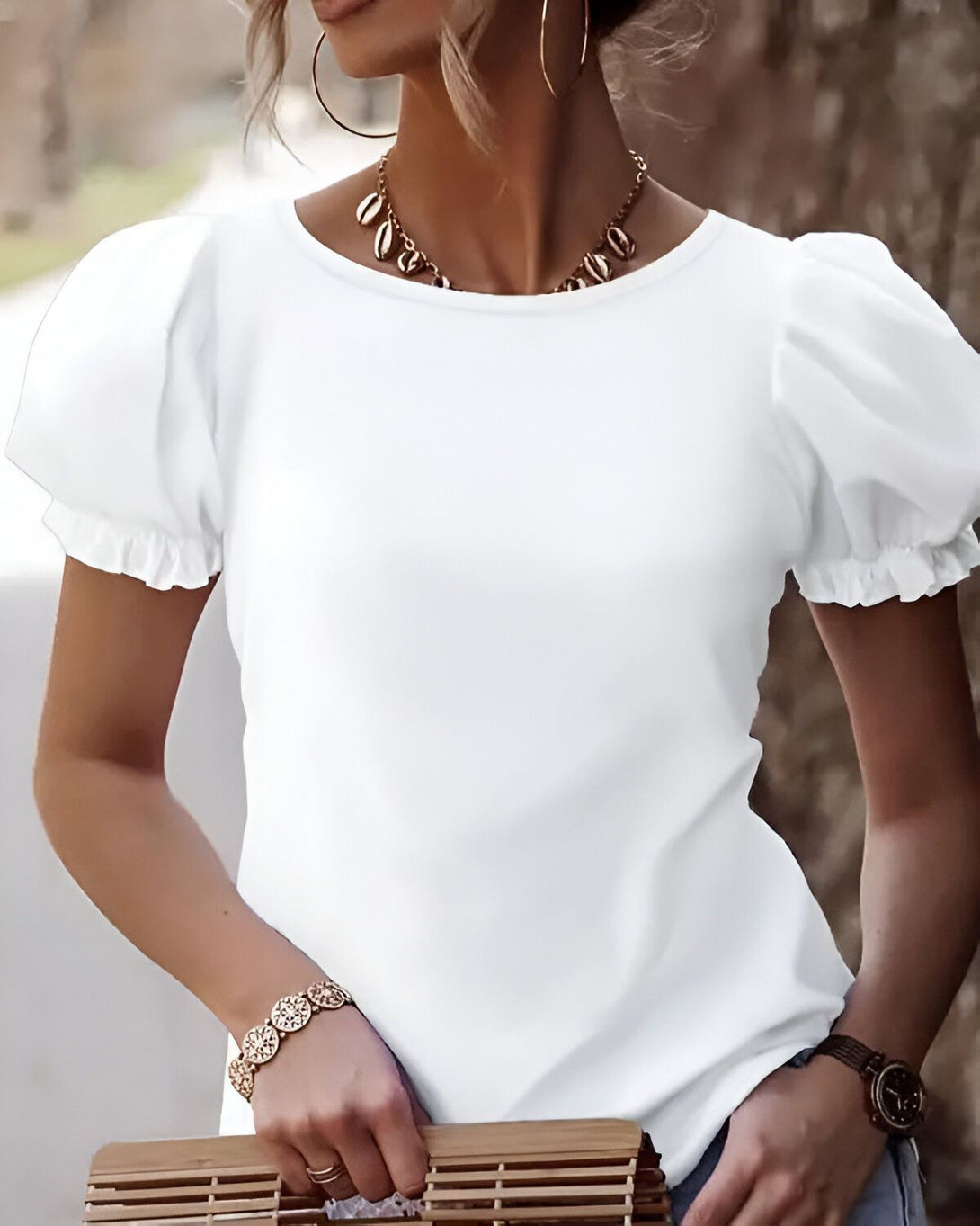 Une jeune femme porte un t-shirt blanc à manches bouffantes et un jean bleu. Elle a les cheveux longs et blonds, et elle porte un collier et des boucles d'oreilles. Elle est debout dans la rue, et elle a l'air heureuse et détendue.