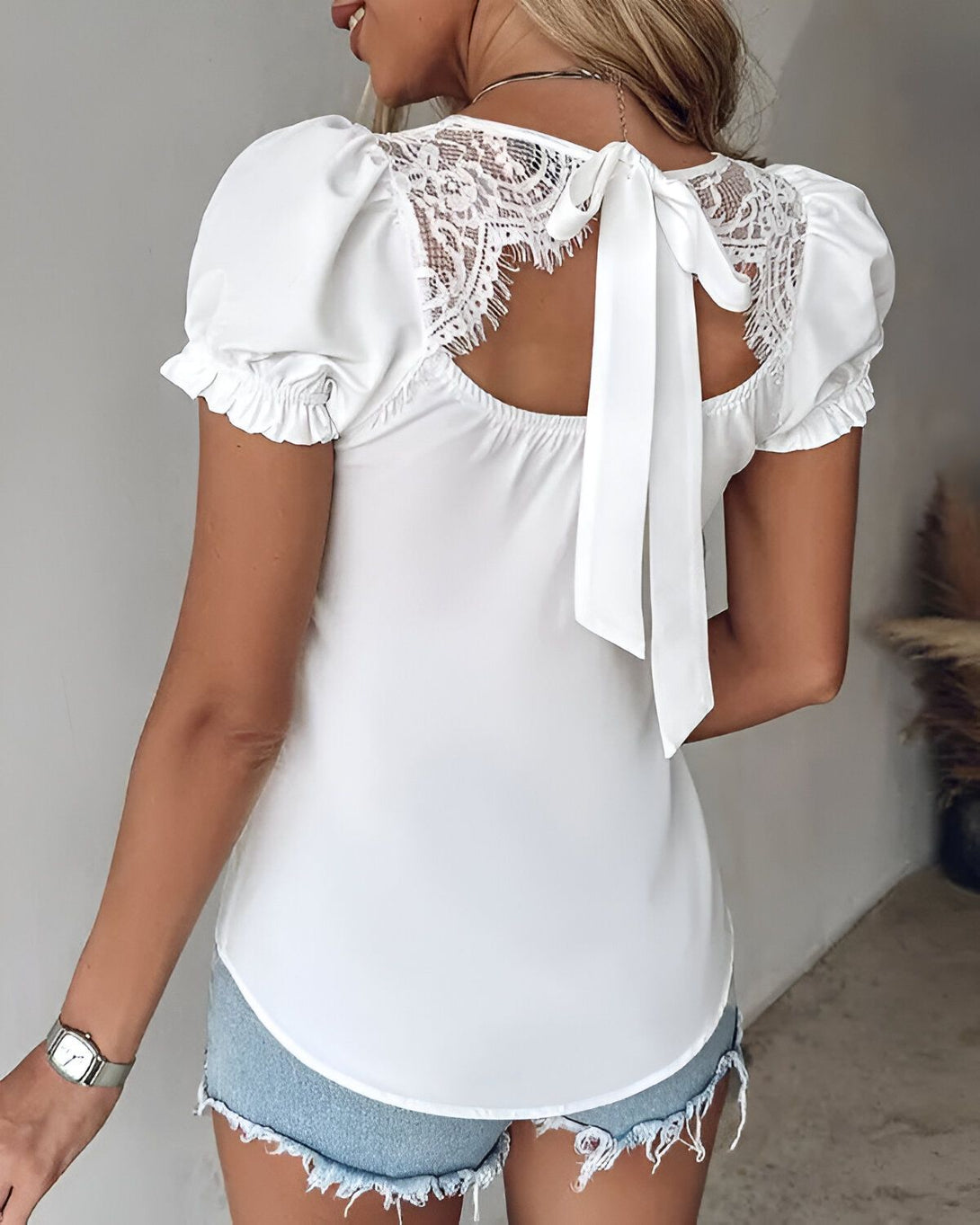 Une femme porte un chemisier blanc avec des manches bouffantes et un dos en dentelle. Le chemisier est rentré dans un short en jean bleu.