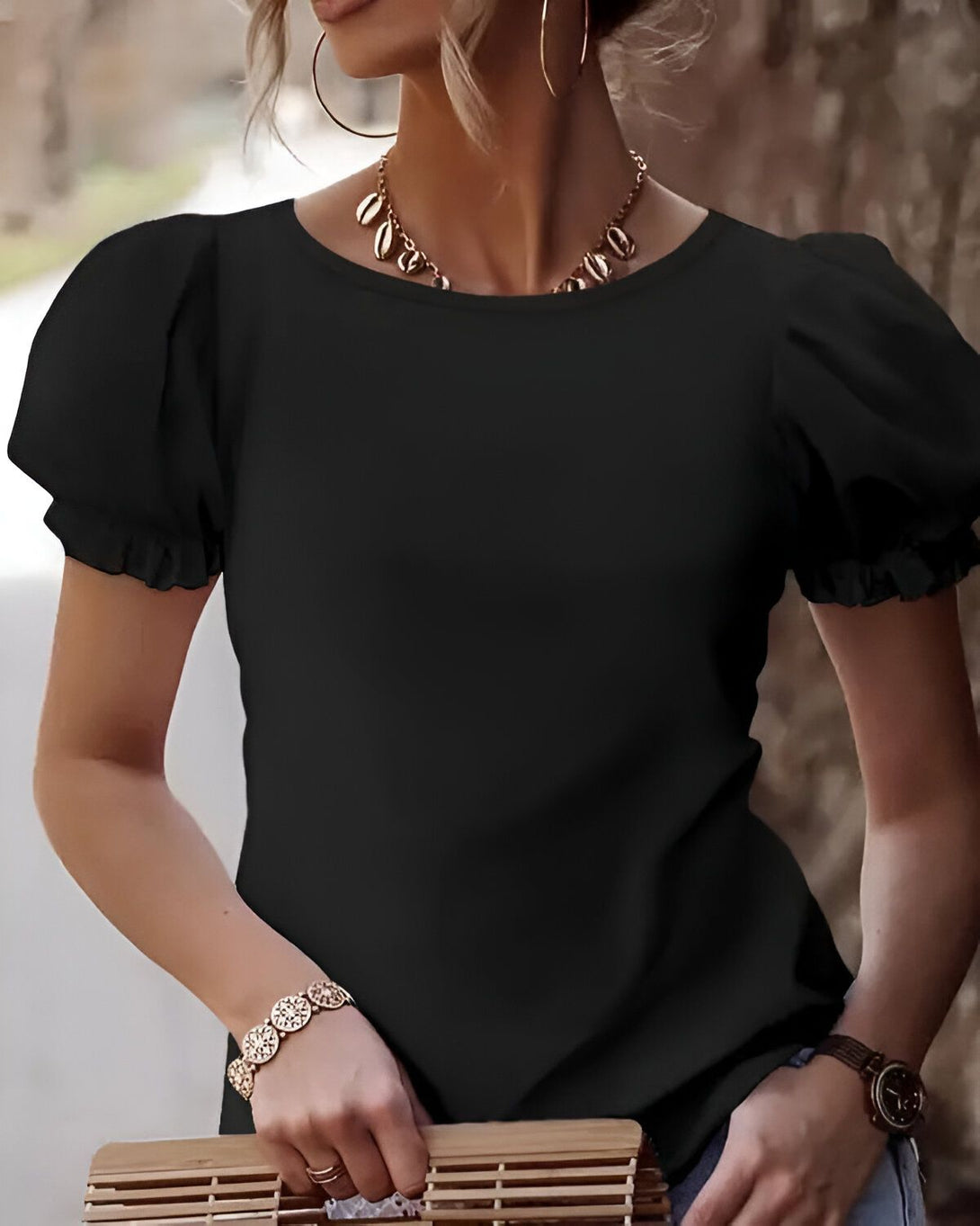 Une femme porte un t-shirt noir à manches bouffantes et un jean. Elle a un collier de coquillages et une montre. Elle porte un sac à main en osier à la main.