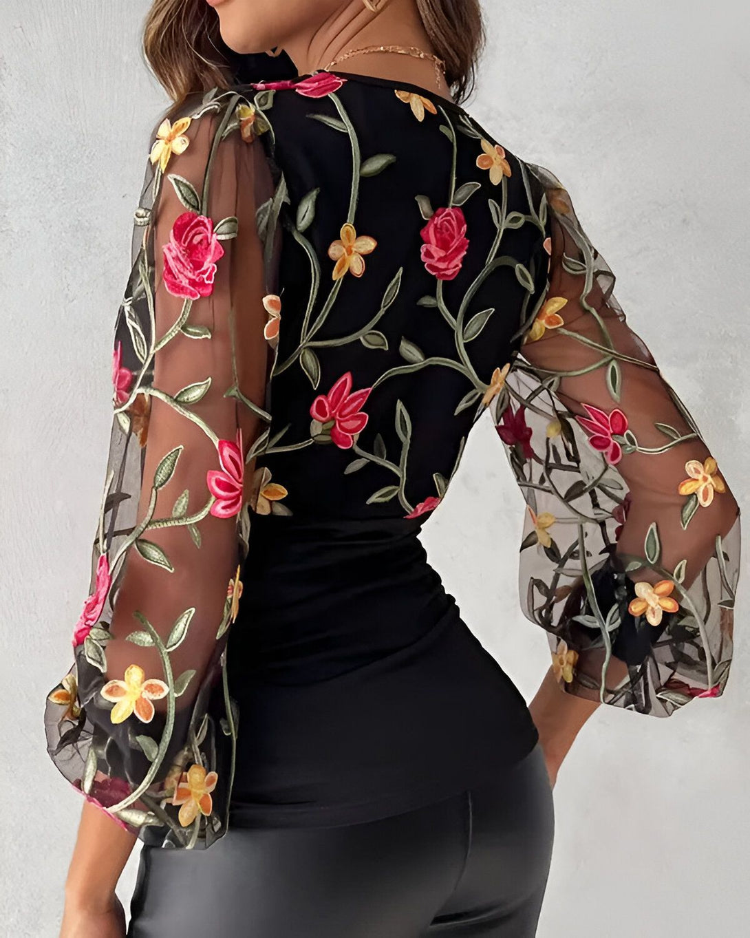 Une femme porte un chemisier noir transparent brodé de fleurs rouges, jaunes et vertes. Le chemisier a des manches bouffantes et un col montant.