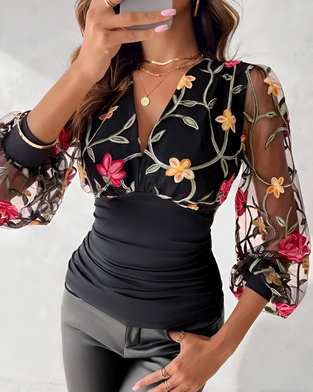 Une femme porte un chemisier noir avec des fleurs brodées. Le chemisier est décolleté en V et a des manches longues. La femme porte également un pantalon noir et des talons noirs.