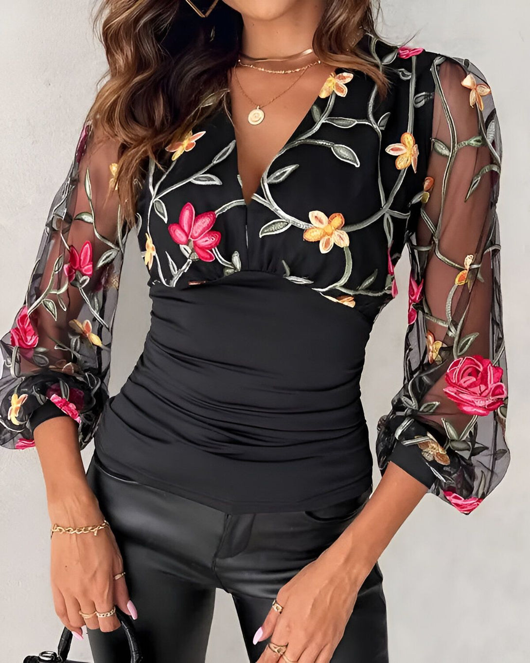 Une jeune femme porte un chemisier noir avec des fleurs brodées. Le chemisier est décolleté en V et a des manches longues. La femme porte également un pantalon en cuir noir et des talons hauts noirs.