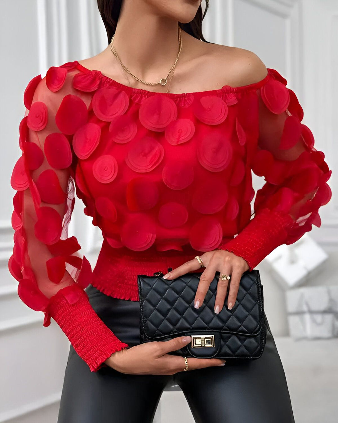 Une femme porte un chemisier rouge à épaules dénudées. Le chemisier est en mousseline de soie et présente un motif de cercles rouges. La femme porte également un pantalon noir et un sac à main noir.