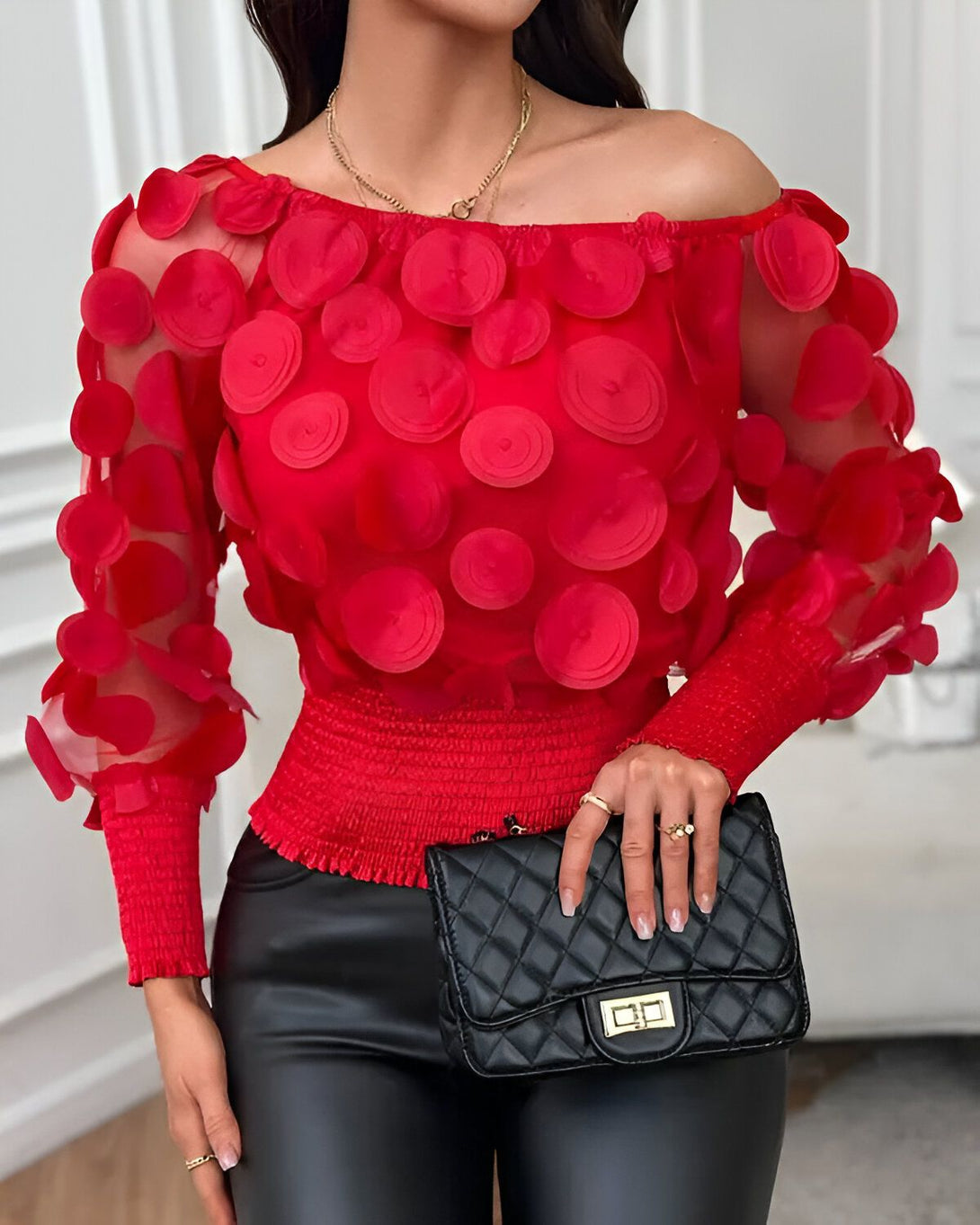 Une femme porte un chemisier rouge à épaules dénudées. Le chemisier est en mousseline de soie et présente un motif de cercles rouges. La femme porte également un pantalon noir et un sac à main noir.