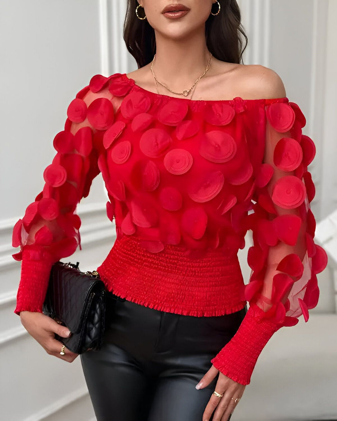 Une femme porte un chemisier rouge à épaules dénudées. Le chemisier est en mousseline de soie et présente un motif de cercles en relief. La femme porte également un pantalon noir et un collier doré.