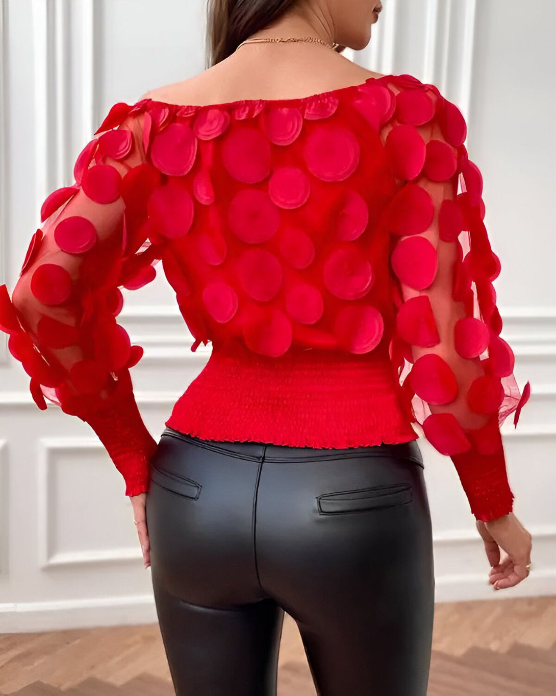 Une femme porte un chemisier rouge à pois et un pantalon en cuir noir. Le chemisier est en mousseline de soie et présente un col bateau ainsi que des manches bouffantes. Le pantalon est en cuir noir et possède une taille haute. La femme est vue de dos.