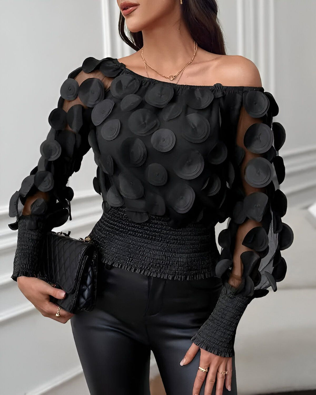 Une femme porte un chemisier noir à épaules dénudées. Le chemisier est orné de cercles noirs en relief. La femme porte également un pantalon noir et un collier doré.