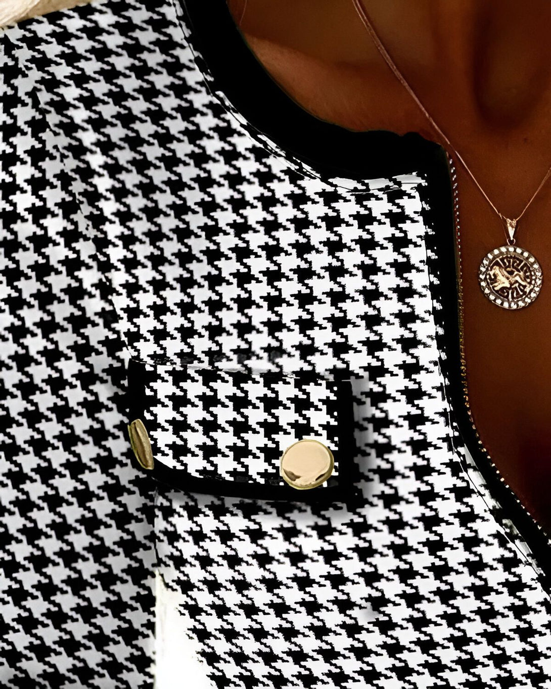 Une femme porte une veste noire et blanche à motif pied-de-poule. La veste est ouverte et on peut voir un chemisier noir en dessous. La femme porte également un collier doré avec un pendentif rond.
