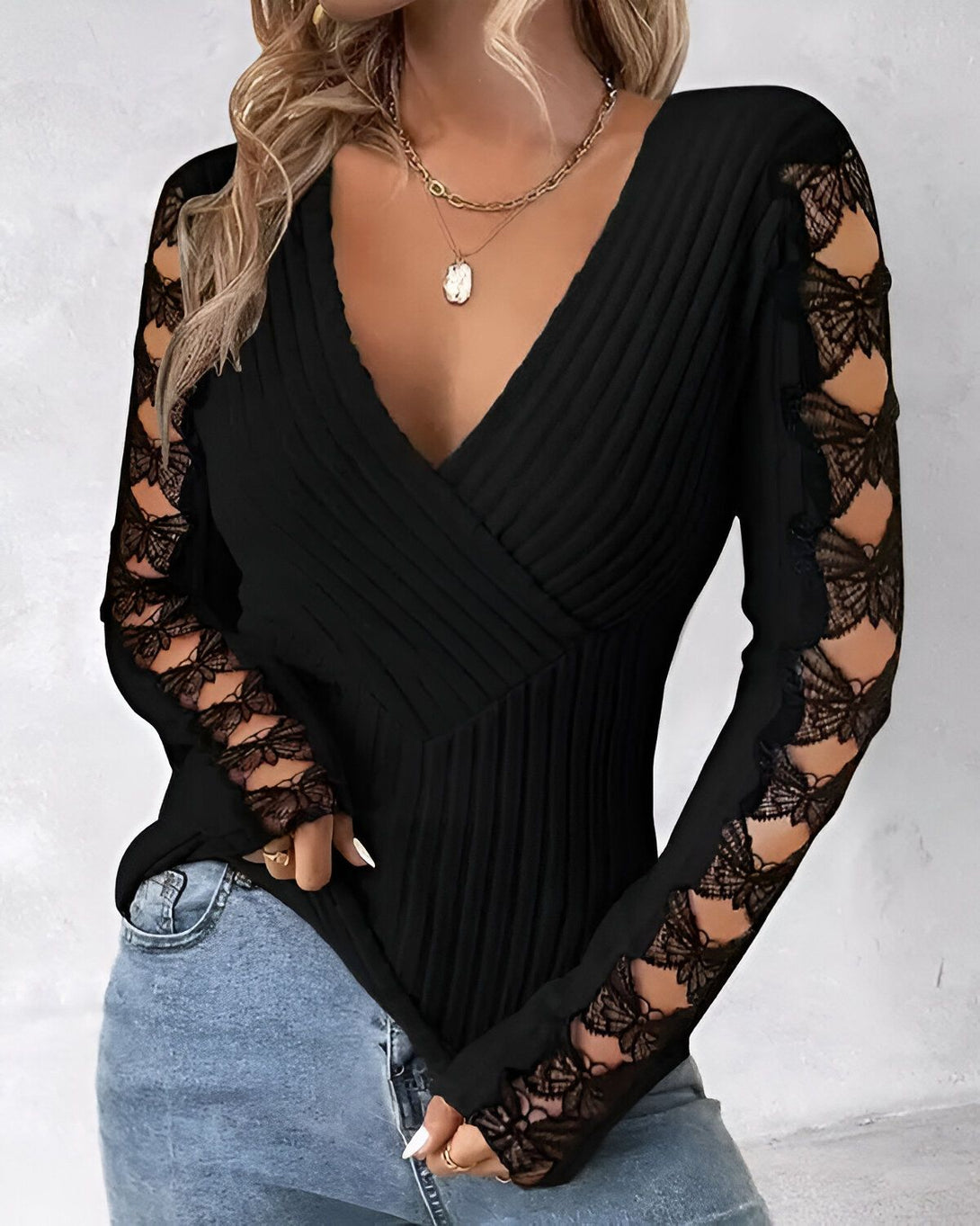 Une femme porte un chemisier noir à manches longues en dentelle. Le chemisier est décolleté en V et cintré à la taille. La femme porte un jean bleu et un collier.