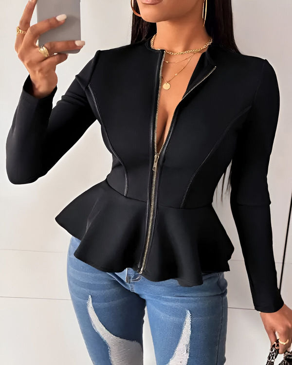 Une femme porte une veste noire à basque et un jean bleu déchiré. Elle a les cheveux longs et noirs et porte du maquillage. Elle est debout dans une pièce blanche et tient un téléphone portable à la main.