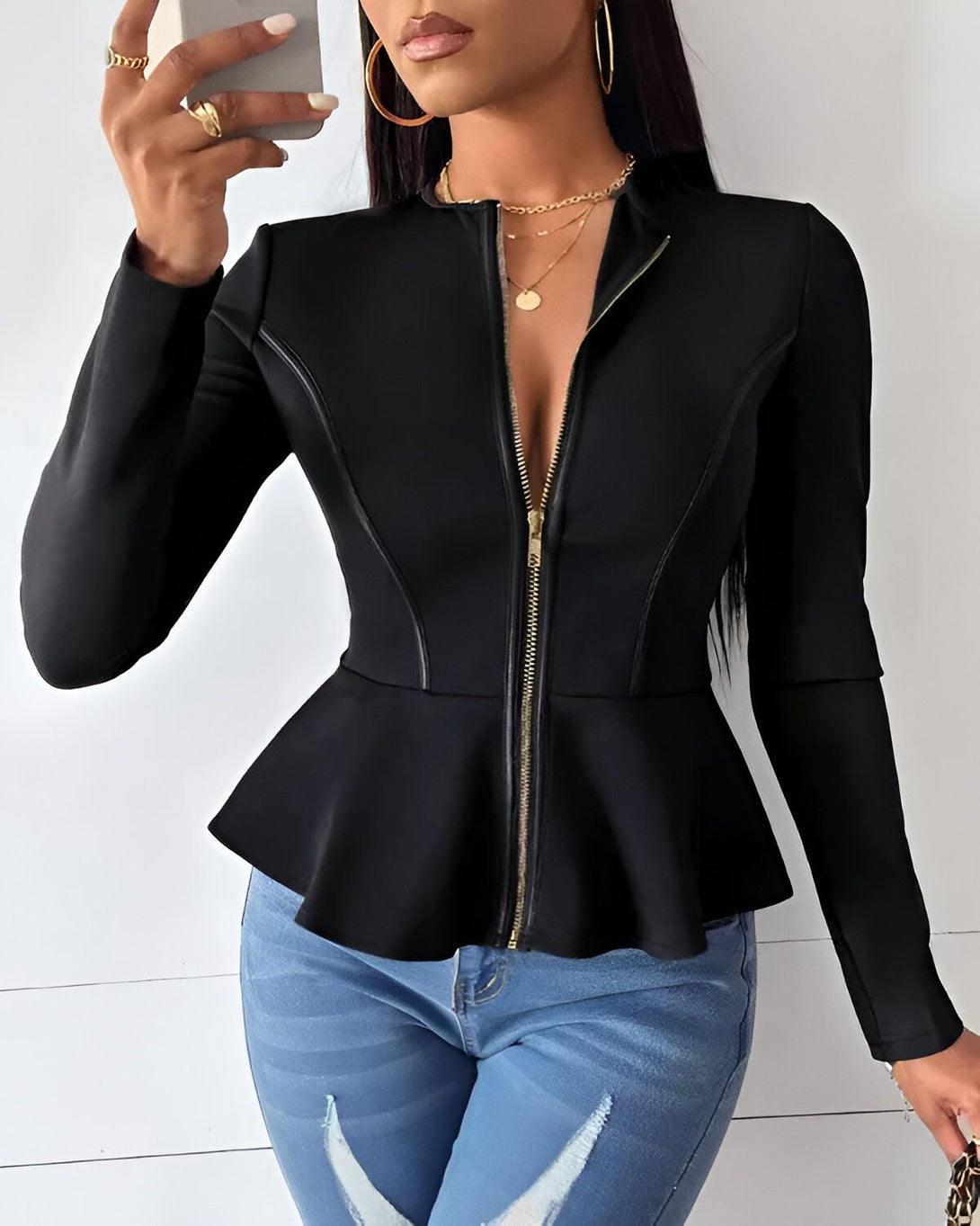 Une femme porte une veste noire cintrée avec un zip sur le devant et des volants au niveau de la taille. Elle porte également un jean bleu déchiré et des talons noirs.