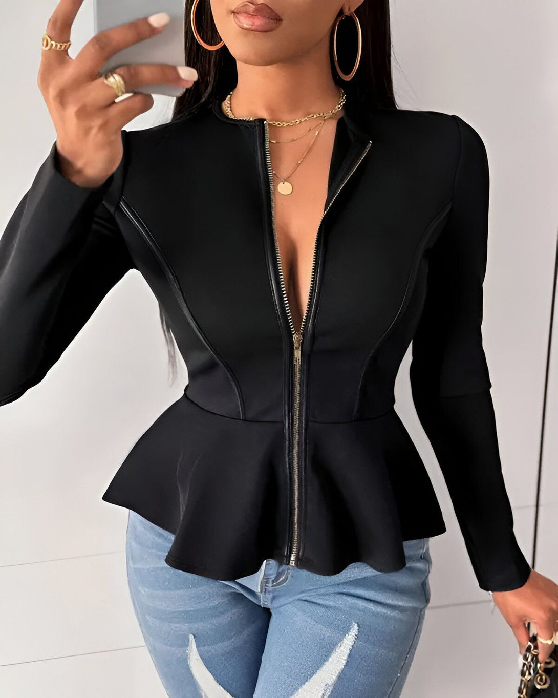 Une femme porte une veste noire à basque avec un zip sur le devant. Elle a les cheveux longs et noirs et porte des boucles d'oreilles rondes. Elle est également maquillée avec du rouge à lèvres et du fard à paupières.