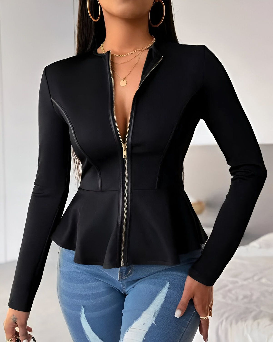 Une femme porte une veste noire cintrée avec un zip doré sur le devant. La veste est évasée au niveau de la taille et possède un col montant. Elle porte également un jean bleu déchiré et des talons noirs.