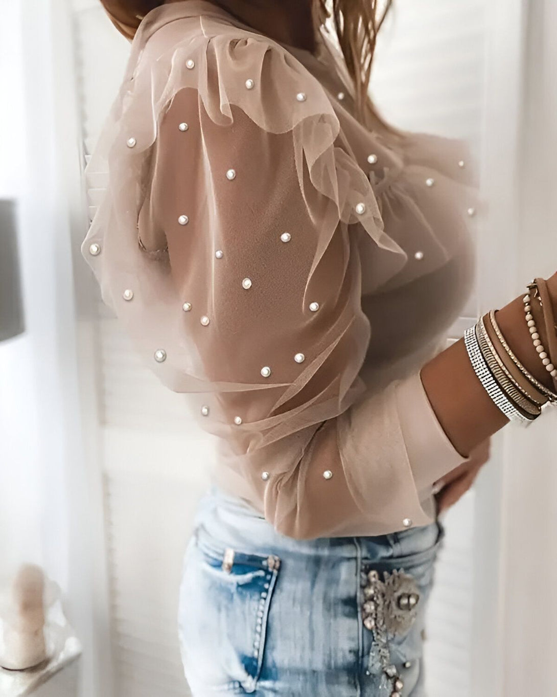 Une femme porte un chemisier beige transparent à pois blancs. Le chemisier est rentré dans un jean bleu. La femme porte également des bracelets et un collier.