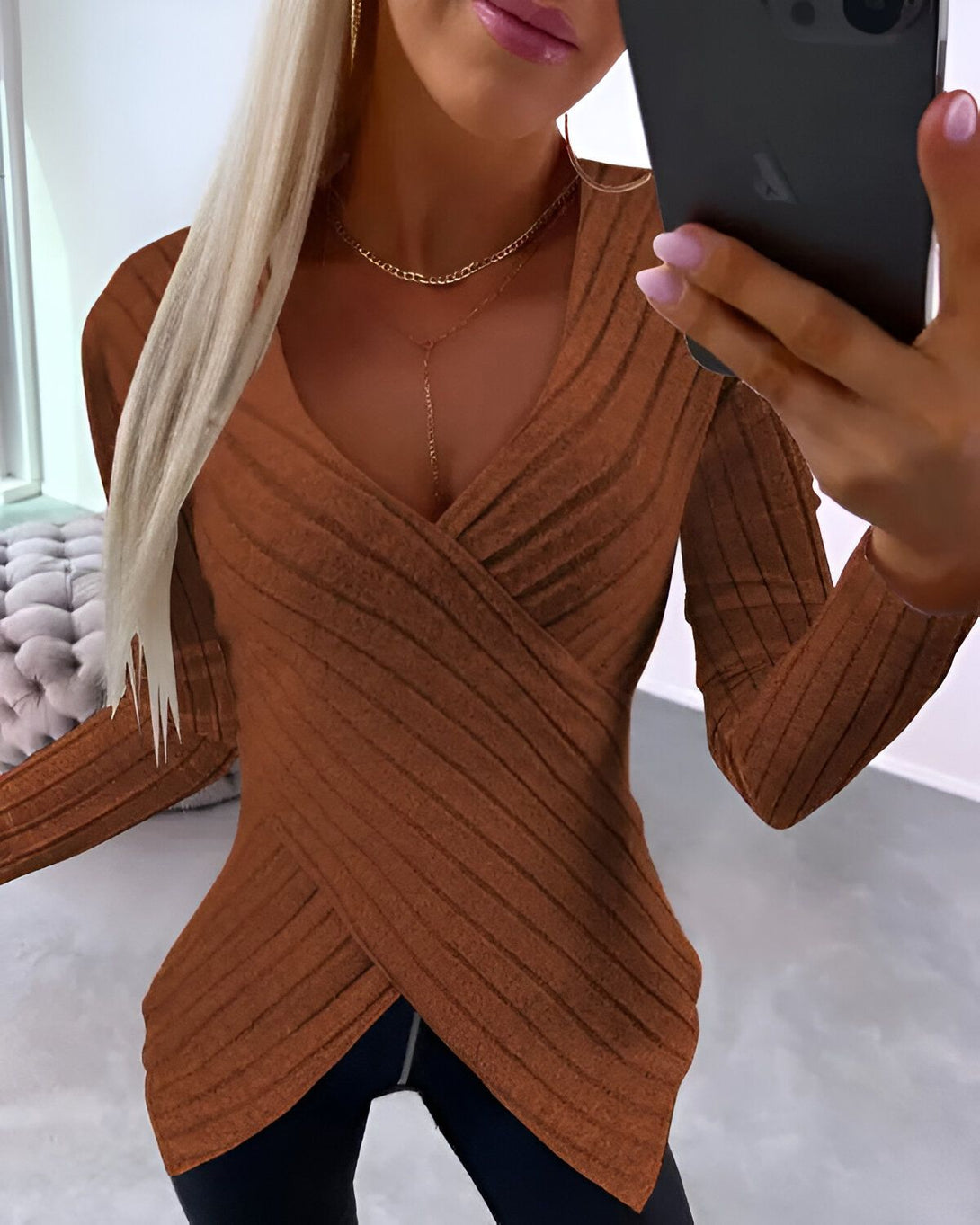 Une femme porte un haut marron à manches longues et décolleté en V. Le haut est croisé sur le devant et rentré dans un pantalon noir. La femme a les cheveux blonds et porte un collier. Elle est debout dans une pièce et tient un téléphone portable à la main.