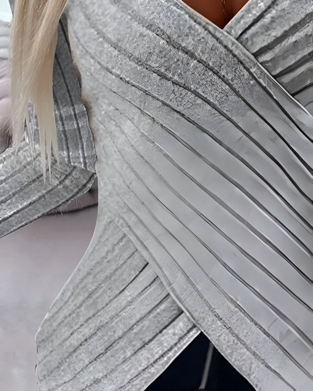 Une femme porte un chemisier gris métallisé à manches longues. Le chemisier est croisé sur le devant et a un col en V. La femme a les cheveux blonds et porte un collier doré.