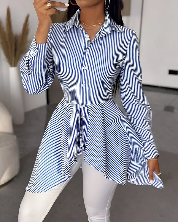 Une femme porte une chemise bleue et blanche à rayures avec un col montant et des manches longues. La chemise est cintrée à la taille et évasée au niveau des hanches. Elle porte un pantalon blanc et des escarpins noirs.