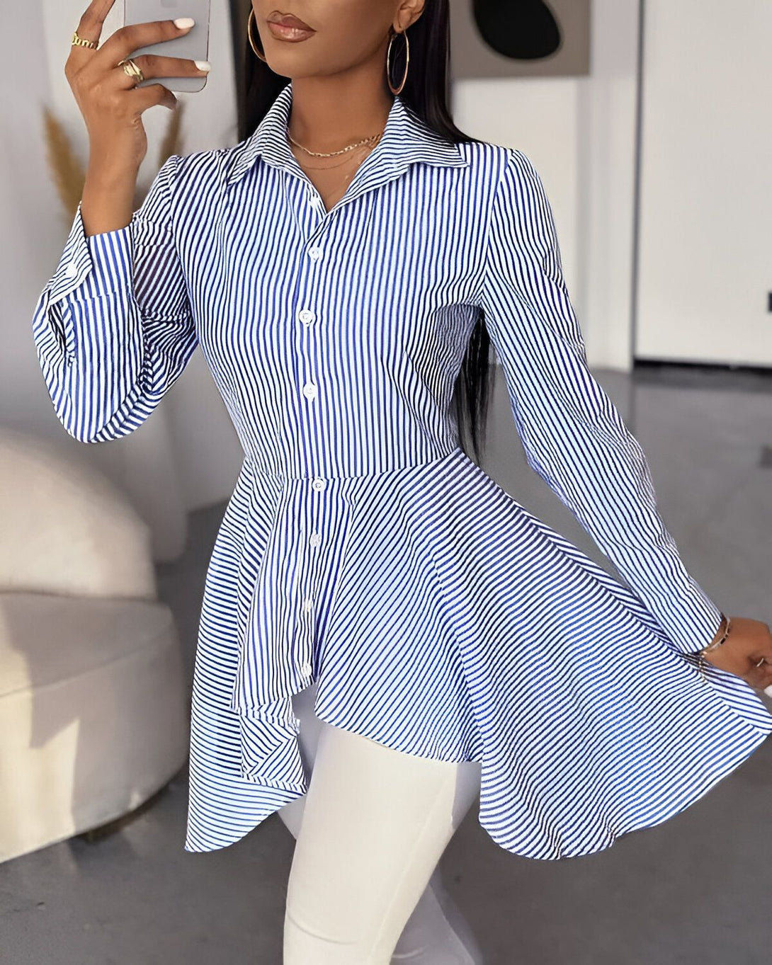 Une femme porte une chemise bleue et blanche à rayures avec un col montant et des manches longues. La chemise est ample et évasée au niveau des hanches. Elle est rentrée dans un pantalon blanc. La femme est debout, elle a une main sur la hanche et l'autre tient un téléphone portable.