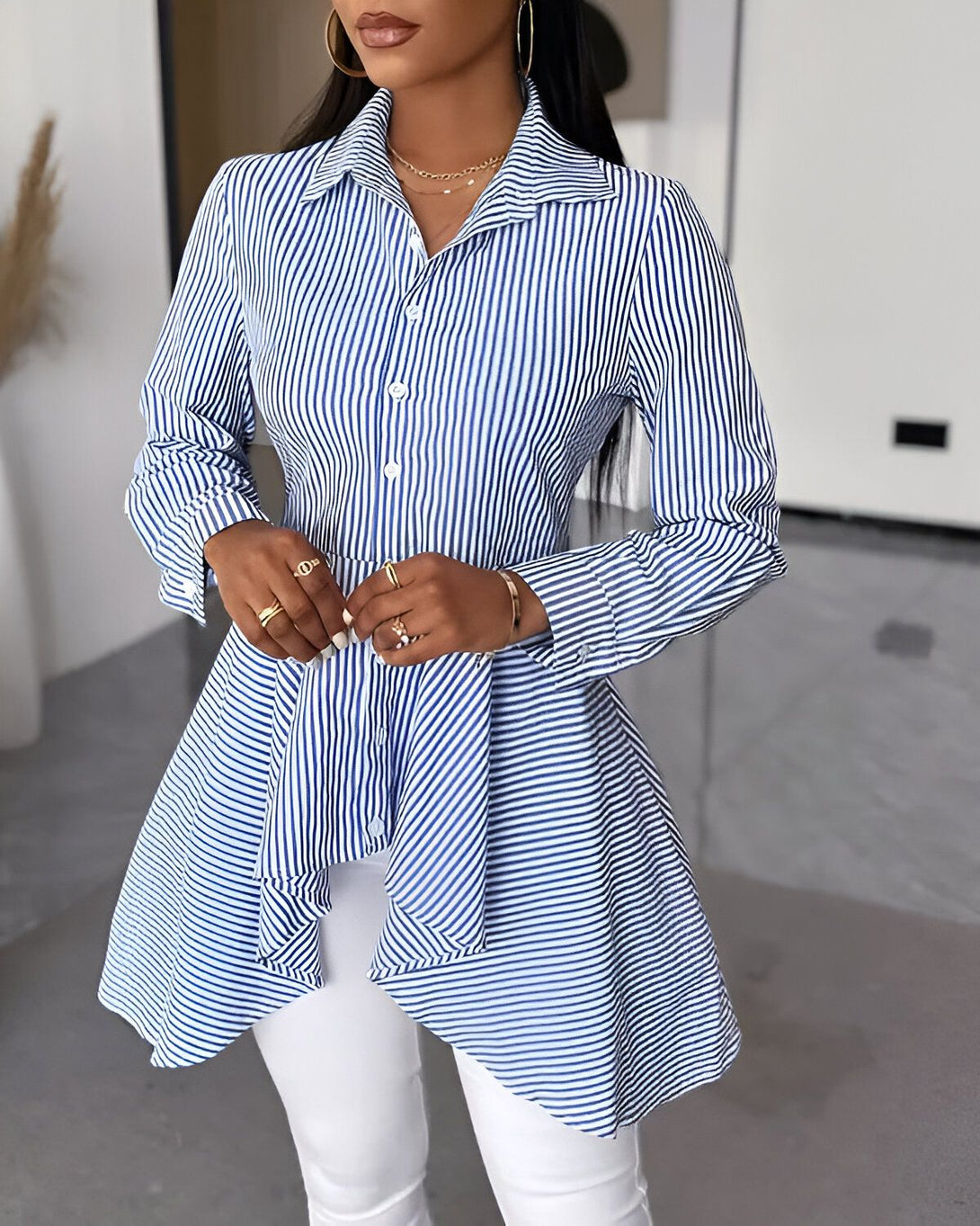 Une femme porte une chemise bleue et blanche à rayures verticales. La chemise est ouverte et on peut voir un soutien-gorge noir. Elle porte également un pantalon blanc et des talons noirs.