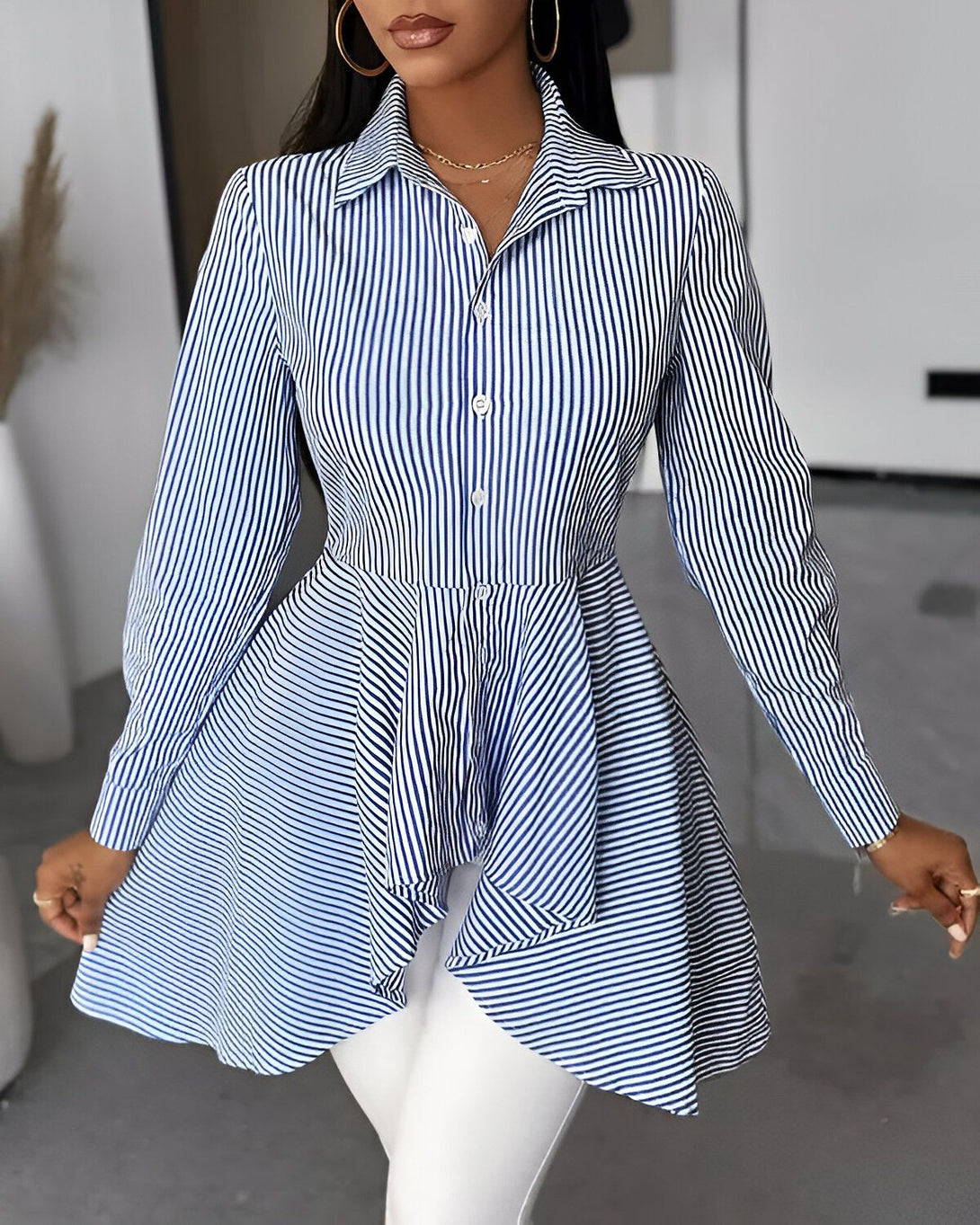 Une femme porte une chemise bleue et blanche à rayures avec un col montant et des manches longues. La chemise est cintrée à la taille et évasée au niveau des hanches. Elle est associée à un pantalon blanc.