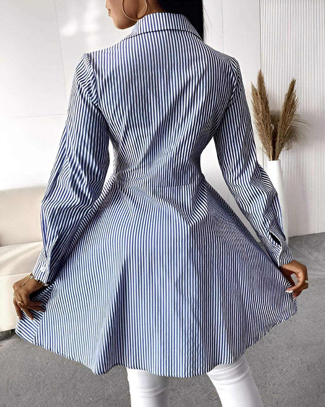 Une femme porte une robe chemise bleue et blanche à rayures verticales. La robe est cintrée à la taille et évasée en bas. Elle a des manches longues et un col montant. La femme est vue de dos.
