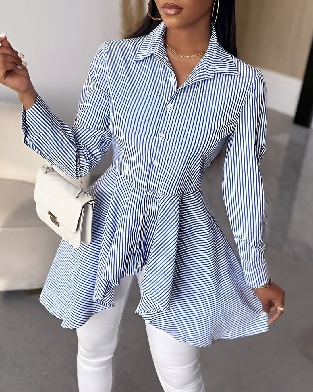 Une femme porte une chemise bleue et blanche à rayures avec un col montant et des manches longues. La chemise est cintrée à la taille et évasée au niveau des hanches. Elle porte un pantalon blanc et des escarpins blancs.