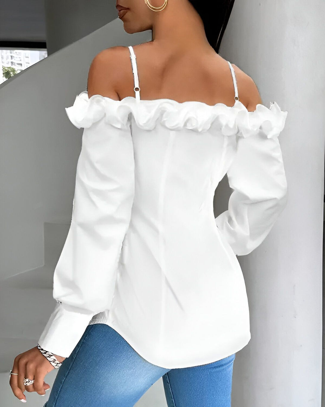 Une femme porte un chemisier blanc à épaules dénudées. Le chemisier est froncé au niveau des épaules et des poignets. Il est rentré dans un jean bleu. La femme est vue de dos.