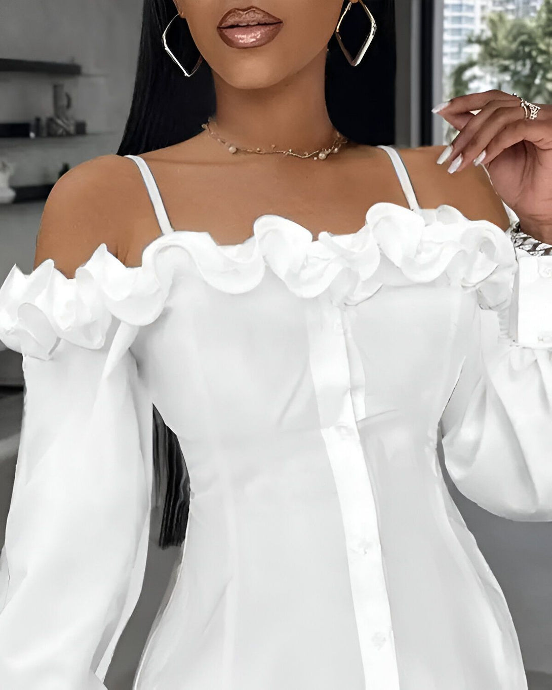 Une femme porte un chemisier blanc à épaules dénudées. Le chemisier est en mousseline de soie et comporte des volants sur les épaules. La femme a les cheveux longs et noirs et porte un maquillage léger. Elle est debout dans une pièce blanche et regarde l'appareil photo.