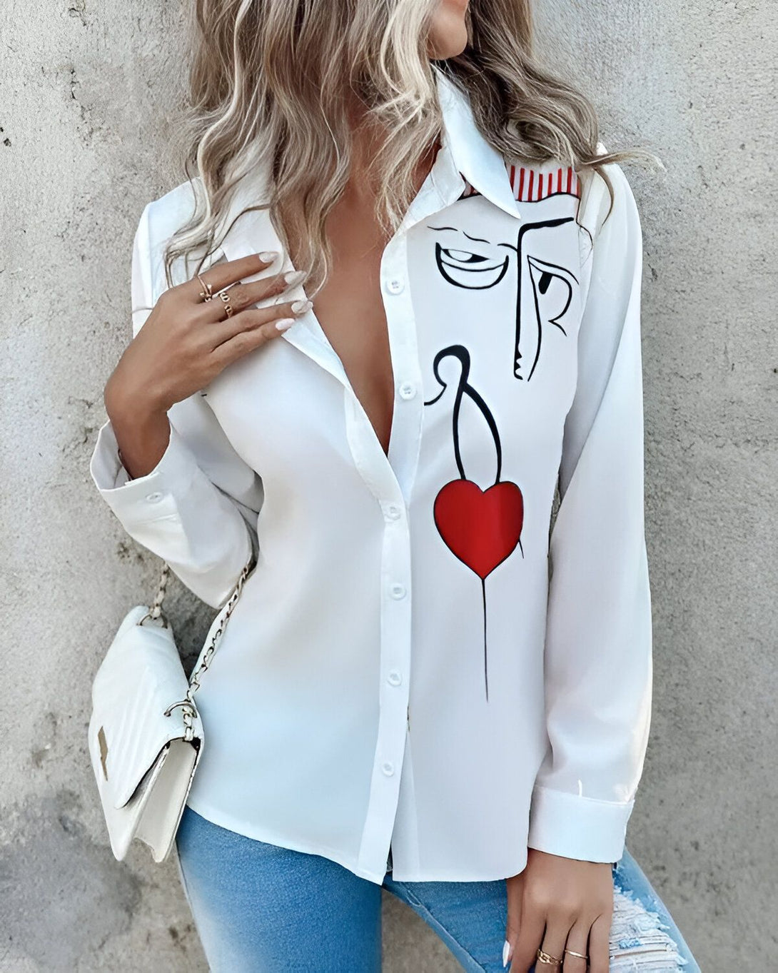 Une femme porte une chemise blanche avec un dessin d'un visage et d'un cœur rouge. La femme est blonde et porte un jean bleu. Elle a un sac à main blanc à la main.