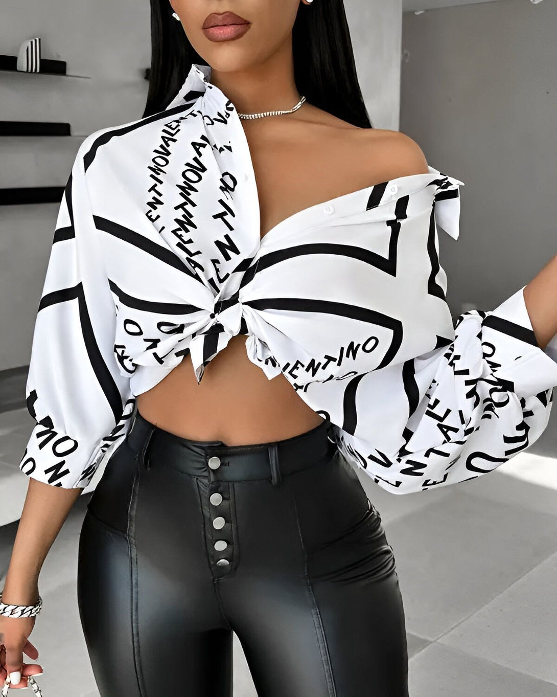 Une femme porte une chemise blanche et noire avec un pantalon en cuir noir. La chemise est ouverte et nouée au niveau du ventre. Le pantalon est taille haute et moulant. La femme est debout, elle a les cheveux noirs et un maquillage léger.