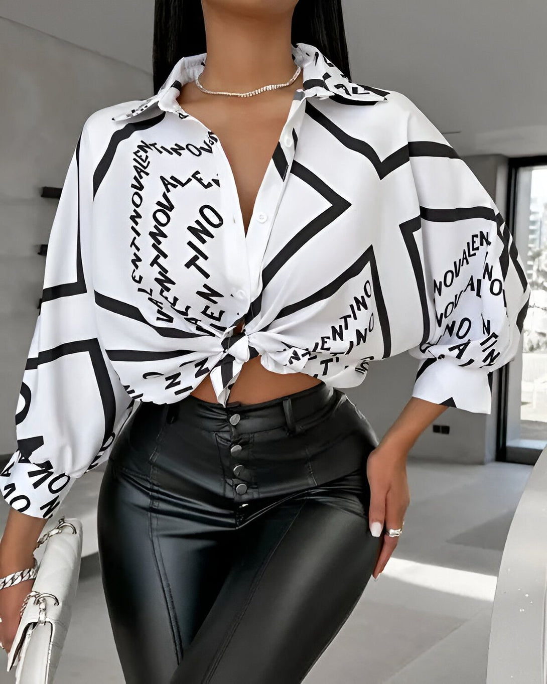 Une femme porte une chemise blanche et noire avec un pantalon en cuir noir. La chemise est ouverte et nouée au niveau de la taille. La femme est debout et porte un sac à main blanc.