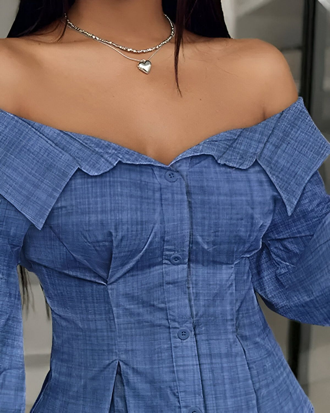 Une femme porte un chemisier bleu à épaules dénudées. Le chemisier est boutonné sur le devant et a un col montant. La femme porte également un collier avec un pendentif en forme de cœur.