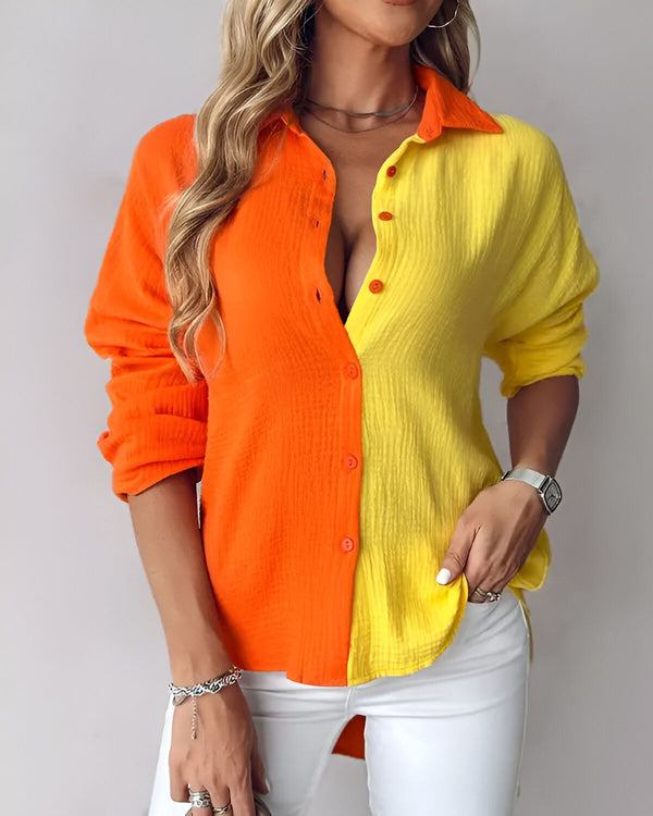 Une femme porte une chemise orange et jaune avec un col et des boutons. La chemise est ouverte sur la poitrine, montrant un décolleté. La femme a les cheveux longs et blonds et porte un pantalon blanc.