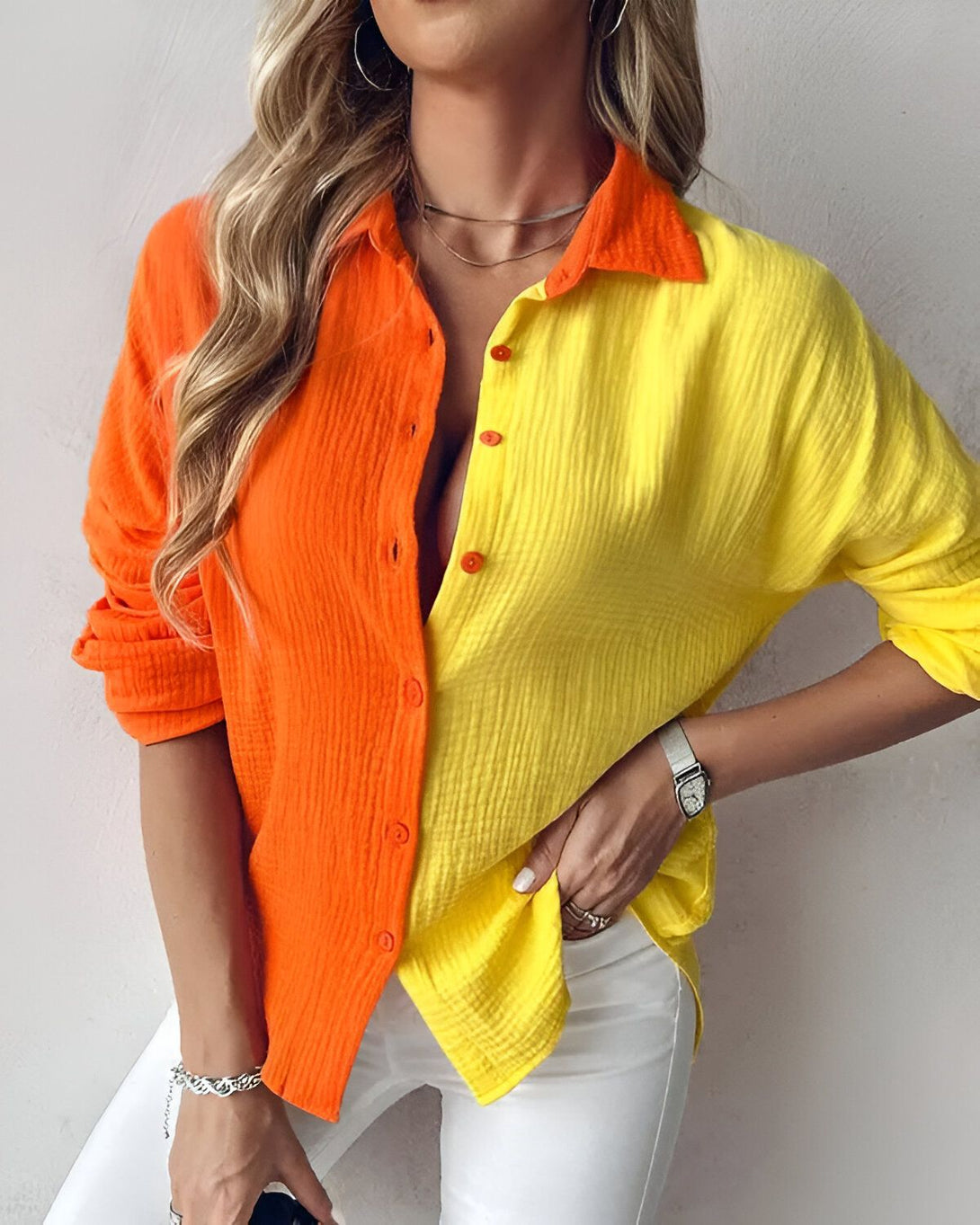 Une jeune femme porte une chemise orange et jaune. La chemise est ouverte et on peut voir son décolleté. Elle porte un pantalon blanc et un collier. Elle a les cheveux longs et blonds. Elle est souriante.