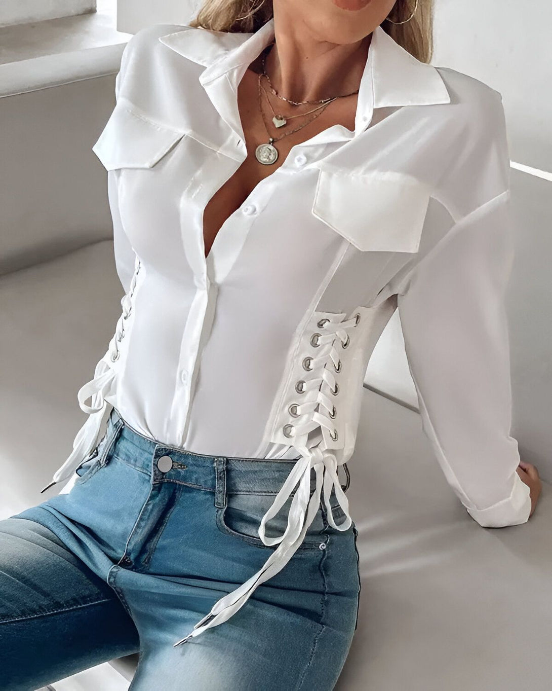 Une femme porte une chemise blanche cintrée avec des lacets sur les côtés. Elle est associée à un jean bleu.