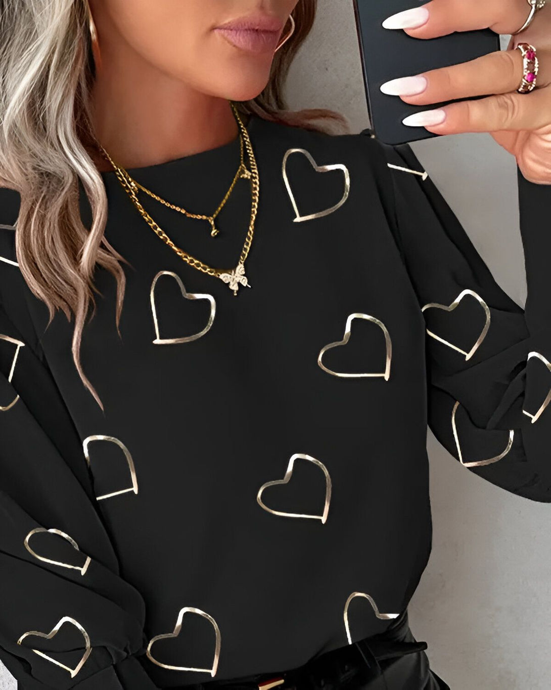 Une femme porte un chemisier noir à manches longues avec des cœurs dorés imprimés. Elle porte également un collier avec un pendentif en forme de papillon.