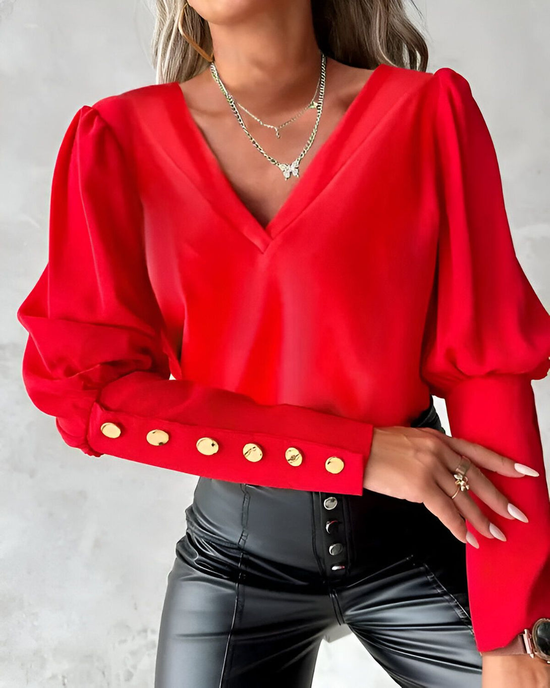 Une femme porte un chemisier rouge à col en V et à manches bouffantes. Le chemisier est fermé par des boutons dorés. La femme porte également un pantalon en cuir noir et un collier avec un pendentif papillon.