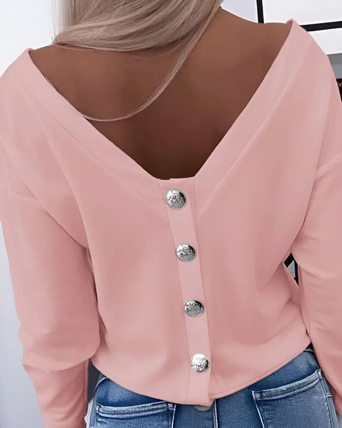 Une femme porte un chemisier rose avec un décolleté en V dans le dos. Le chemisier est boutonné dans le dos.