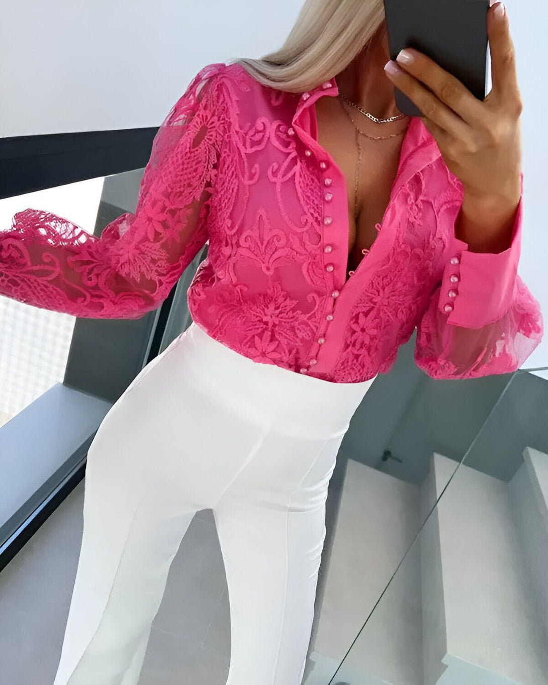 Une femme porte un chemisier rose en dentelle transparente avec des boutons et un pantalon blanc.