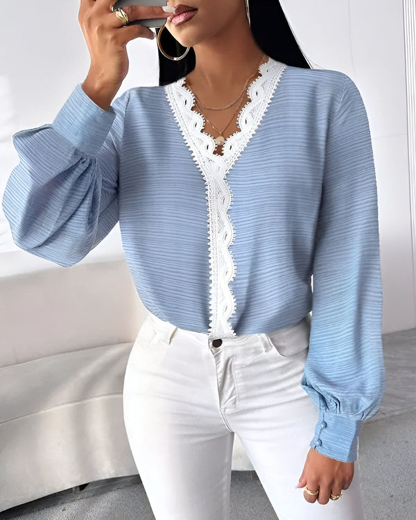 Une femme porte un chemisier bleu avec un col en dentelle blanche. Elle a les cheveux longs et noirs et porte un pantalon blanc. Elle est debout dans un bureau et tient un téléphone portable à la main.