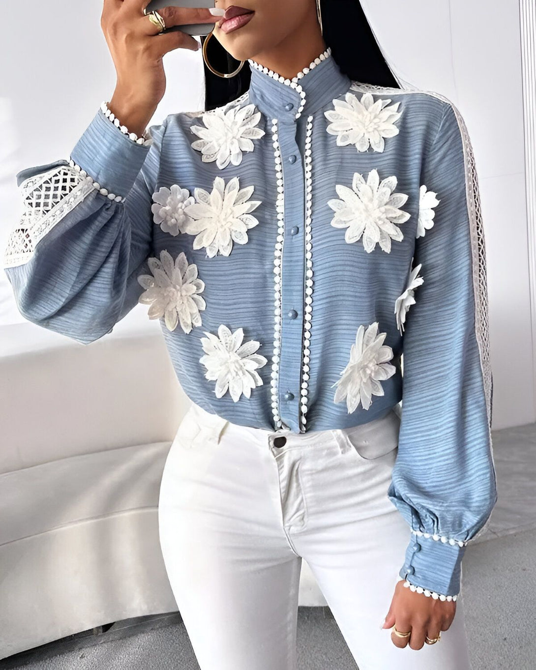 Une femme porte un chemisier bleu avec des fleurs blanches en relief. Le chemisier est fermé par des boutons blancs. La femme a un pantalon blanc et des talons blancs.
