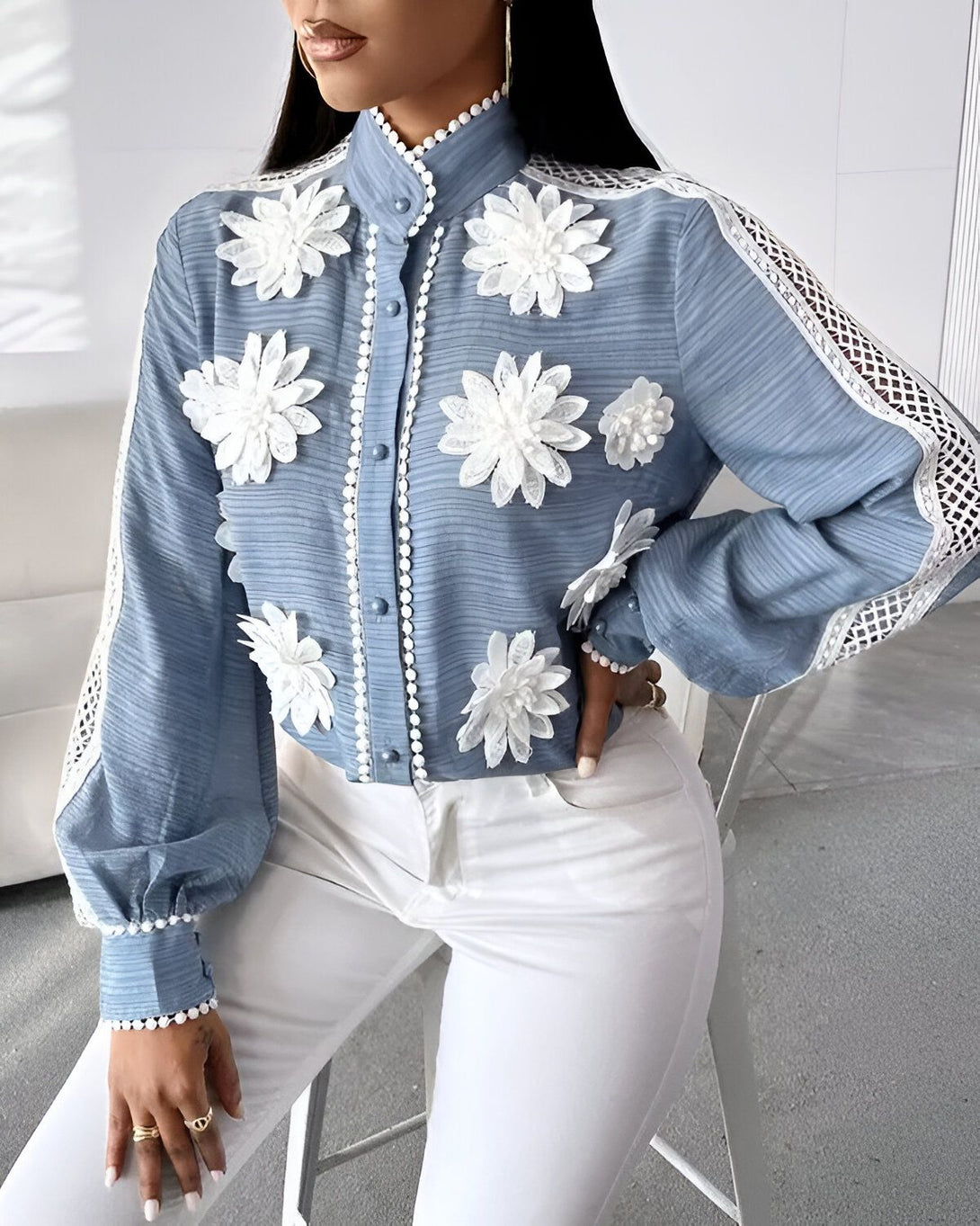 Une femme porte un chemisier bleu avec des fleurs blanches en relief. Le chemisier est fermé par des boutons blancs. La femme porte également un pantalon blanc.