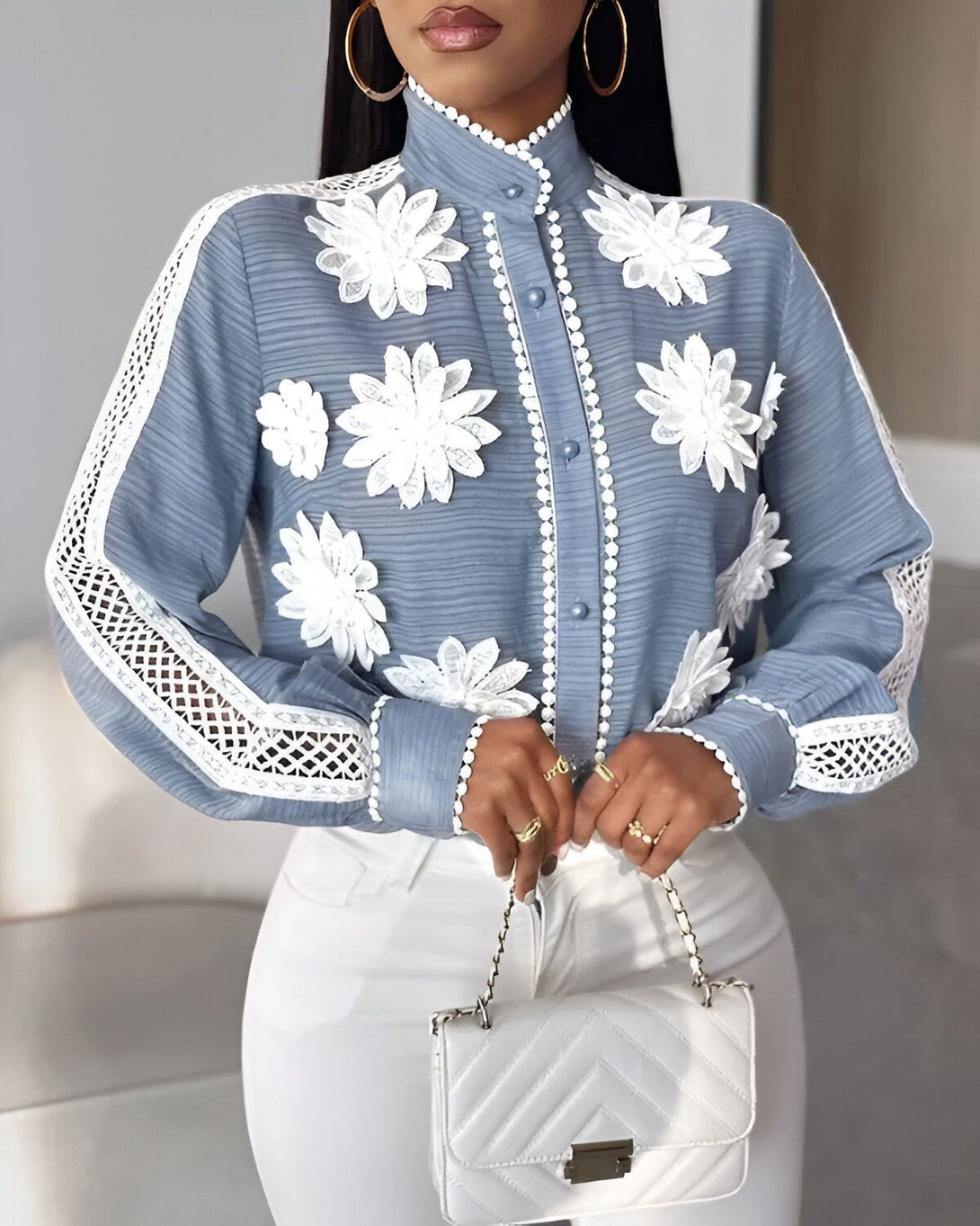 Une femme porte un chemisier bleu avec des fleurs blanches brodées. Le chemisier est fermé par des boutons blancs. La femme porte également un pantalon blanc et un sac à main blanc.