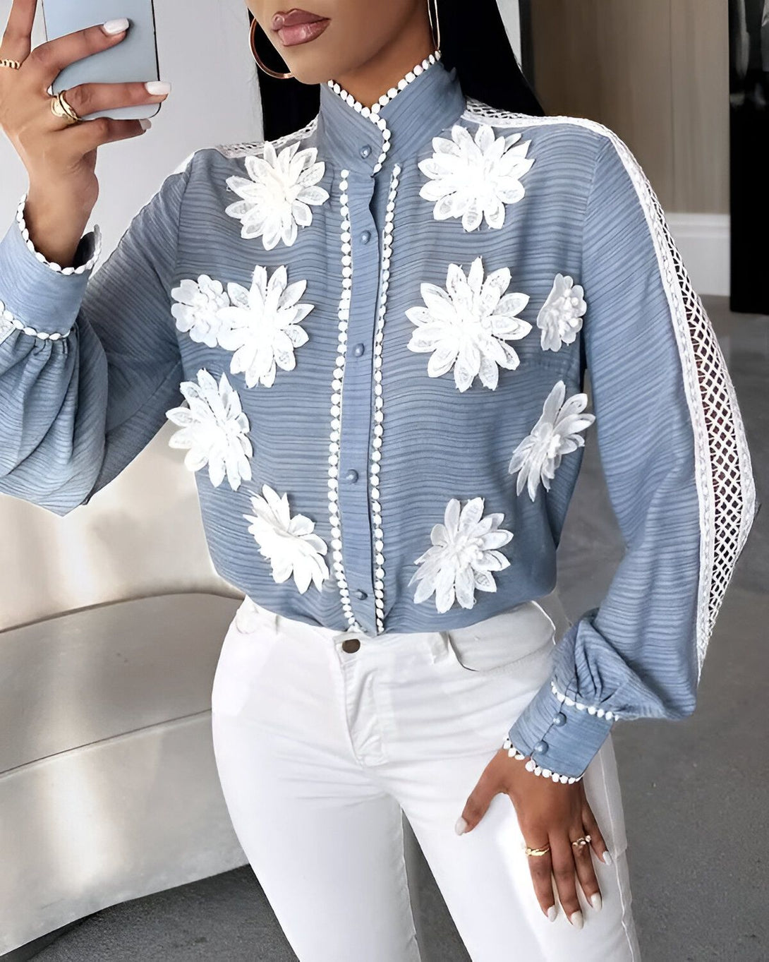 Une femme porte un chemisier bleu avec des fleurs blanches en relief. Le chemisier est fermé par des boutons blancs. La femme porte également un pantalon blanc et des talons hauts.